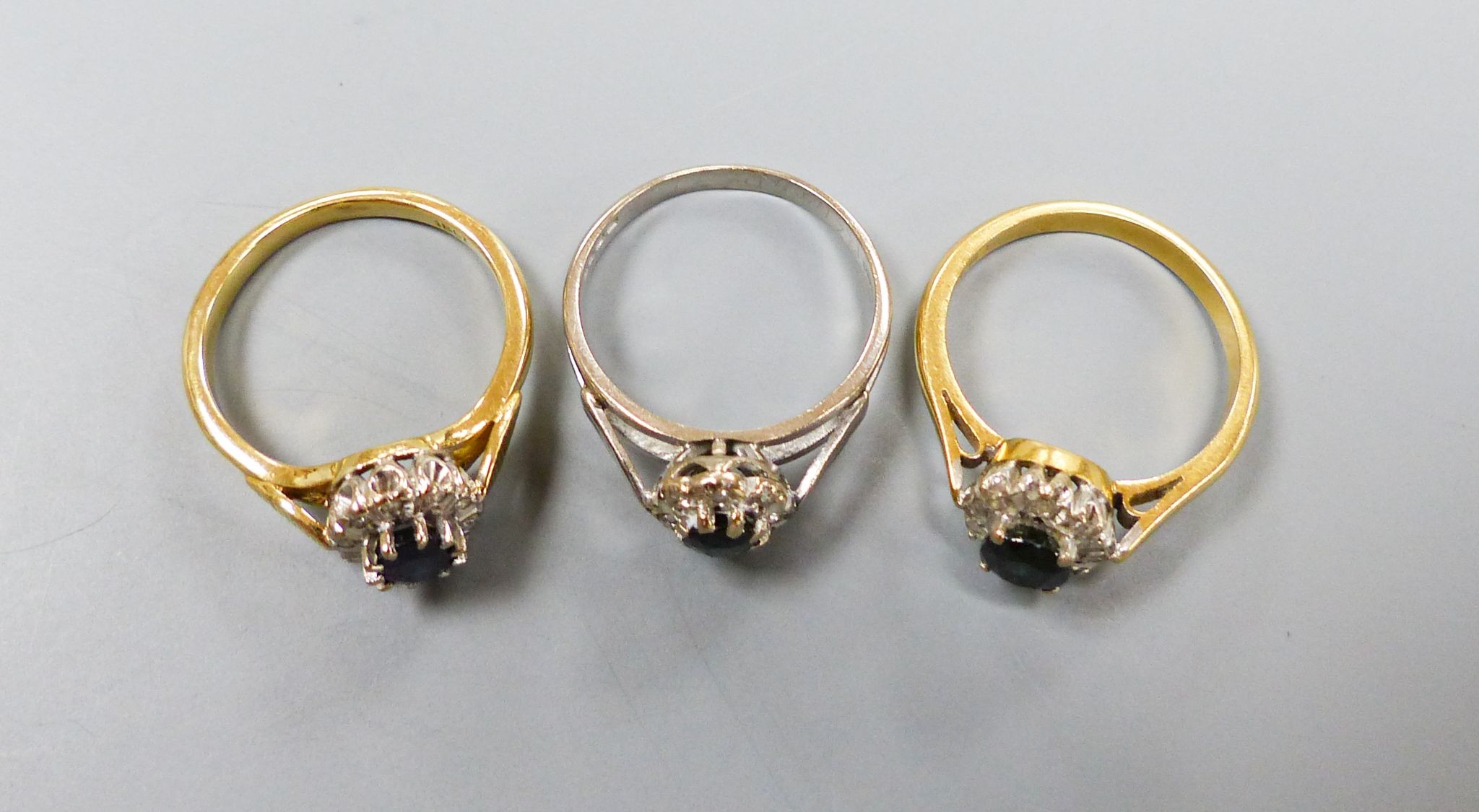 Three modern sapphire and diamond set oval cluster rings, including 18ct white gold, size M and 18ct, size L/M, (gross 7.2 grams and one yellow metal, size L/M, gross 3.8 grams.